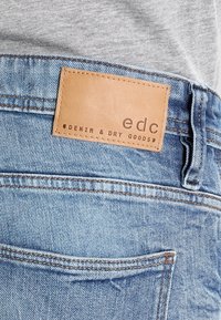 Světle modré džínové kalhoty s texturovanou hnědou koženou etiketou, na které je uvedeno "edc" a "DENIM & DRY GOODS", viditelné švy a detaily kapes.