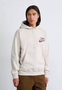 En ung mand med langt, mørkt hår har en lys beige hoodie med et stiliseret Nike-logo og en frontlomme, mod en ensfarvet baggrund.
