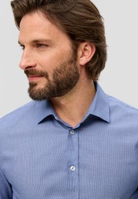 Lichtblauwe shirt met korte mouwen en een gestructureerd patroon, met een button-down kraag en lichte knopen, gedragen door een man met een baard en verfomfaaid haar.