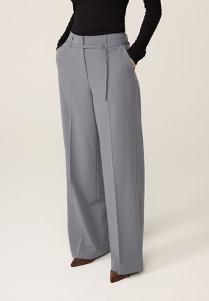 Grijze broek met wijde pijpen en een riem met zijgesp, gedragen met een zwarte top en bruine puntige schoenen, handen in de zakken.