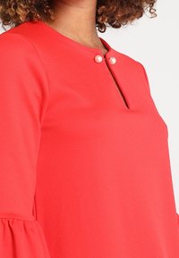 Rote Bluse mit rundem Halsausschnitt, zwei Perlenakzenten und ausgestellten Ärmeln. Der Stoff wirkt glatt mit einer feinen Struktur.