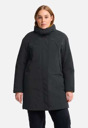 Giovane donna che indossa un cappotto invernale nero di lunghezza media con collo alto e cappuccio, in piedi davanti a uno sfondo chiaro e uniforme.