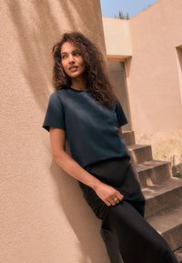 Marineblauw kortärmlig top in een gladde stof, gecombineerd met een zwarte satijnen rok. Weelderig krullend haar en zilveren ringen zijn zichtbaar. Getextureerde beige muur.