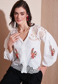 White blouse with floral embroidery in red, yellow, and green. Features lace details, balloon sleeves, and a scalloped hem.