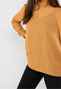 Blusa senape con collo alto, maniche lunghe e polsini arricciati. Presenta un sovrapposto trasparente con piccoli pois verdi.