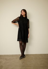 Melna kleita ar mežģīnēm apdrukātām īsām piedurknēm, pogu priekšpusi un viegli izpletotu siluetu. Kopā ar melnām zeķubiksēm un svītrainām, smailām kurpēm.