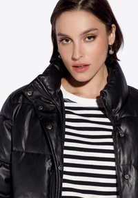 Chaqueta de plumas negra de cuero con cuello alto, textura brillante y botones plateados, superpuesta a una camisa de rayas negra y blanca.