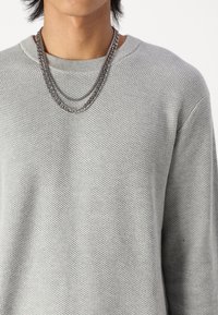 Maglione grigio chiaro a maglia con un motivo testurizzato, dotato di scollo a girocollo e catene d'argento sovrapposte che poggiano sul colletto.
