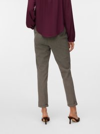 Persona ar sagriestām pelēkām biksēm, bordo garām piedurknēm blūzi un brūnām augstpapēžu kurpēm ar atvērtu aizmuguri, stāv uz neitrāla fona.