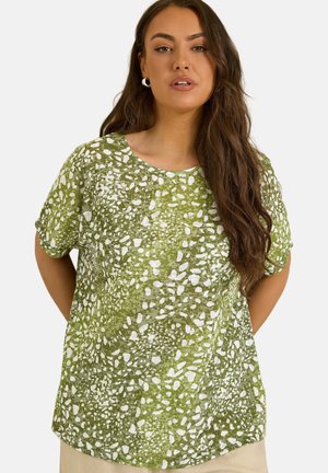 Kobieta z długimi brązowymi włosami, ubrana w zieloną bluzkę z krótkim rękawem w biały abstrakcyjny wzór w kropki, stojąca z rękami za plecami.