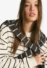 Szürke-fehér csíkos sál krémszínű pulóver fölött. A sál puha, texturált anyagból készült; a pulóveren sötét csíkok találhatók világos alapon.