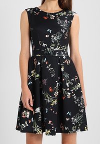 Robe noire sans manches avec motifs floraux multicolores et papillons, taille cintrée et jupe évasée portée par une femme.