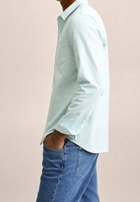 Chemise boutonnée vert menthe clair avec une poche poitrine, manches longues et détails aux poignets, associée à un jean en denim bleu.