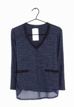 Pull en maille bleu marine à manches longues avec bordure noire au niveau du col en V et des poches, superposé à un tissu bleu marine translucide à l’ourlet, suspendu sur un cintre noir.