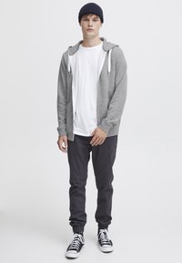 Felpa grigia con cerniera e lacci bianchi sopra una maglietta bianca. Pantaloni scuri e sneakers in bianco e nero completano l'outfit.