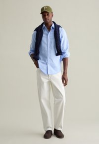 Camisa de botones azul claro, suéter de punto en cable azul marino caído sobre los hombros, pantalones blanco grisáceo y zapatos de ante marrón. Gorra verde oliva con la letra "P".