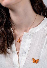 Kuldne kaelakee, millel on oranž liblikapendant, kantud valge tekstuuriga pluusi peal, millel on sobiv liblikapandla pins.