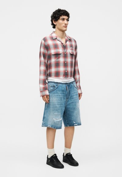 Νέος άνδρας με σγουρά μαλλιά που φοράει κόκκινο και λευκό καρό πουκάμισο, φαρδιά σκισμένα τζιν σορτς, άσπρες κάλτσες και μαύρα αθλητικά παπούτσια, στέκεται μπροστά σε λευκό φόντο.