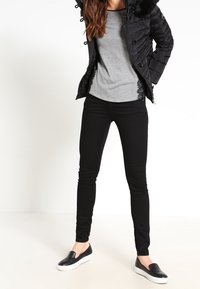 Černá péřová bunda s umělou kožešinou na kapuci, šedé tričko s dlouhým rukávem, černé slim-fit kalhoty a černé nazouvací boty s bílou podrážkou.