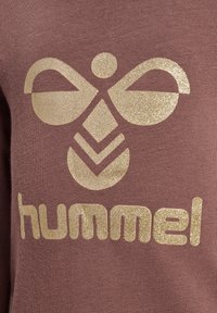 Brązowy dzianinowy sweter z błyszczącym złotym logo przedstawiającym stylizowane kształty motyli oraz słowo "hummel" napisane dużymi literami.