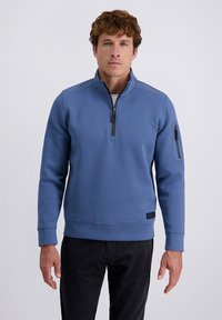 Uomo che indossa un maglione a mezze maniche blu con zip sulla manica sinistra e pantaloni neri, in piedi contro uno sfondo chiaro e semplice.