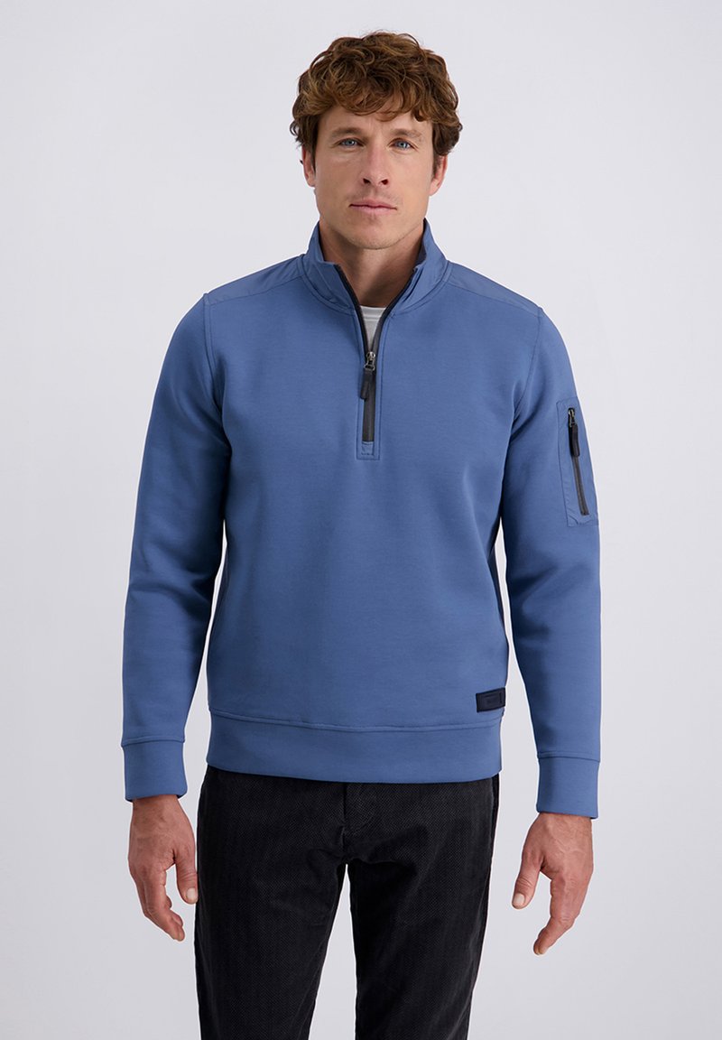 Uomo che indossa un maglione a mezze maniche blu con zip sulla manica sinistra e pantaloni neri, in piedi contro uno sfondo chiaro e semplice.