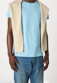 Jasnoniebieska bawełniana koszulka z krótkim rękawem z beżowym swetrem w prążki luźno zarzuconym na ramionach. Ciemnoniebieskie dżinsy. Skupienie na kontrastach tkanin i kolorów.