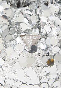 Silver sequin bag featuring circular sequins and a triangular metal logo tag with "GUESS U.S.A." embossed in the center. Reflective texture.