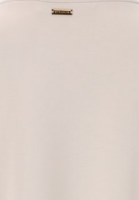 Light beige top made of soft fabric, featuring a smooth texture and a small rectangular gold hardware detail with branding at the neckline.