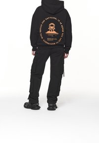 Persona vistiendo pantalones cargo negros y una sudadera con capucha negra con un gráfico de palmera naranja y texto en la parte de atrás, de pie contra un fondo blanco.