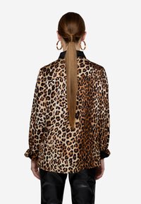 Blusa con stampa leopardata e texture setosa, colletto nero e maniche lunghe, abbinata a pantaloni in pelle. Vestibilità flessibile e motivo vivace.