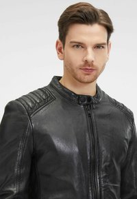 Giacca in pelle nera con zip frontale, chiusura a scatto e accenti imbottiti sulle spalle. Tessuto liscio con design aderente e colletto alto.