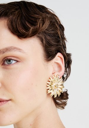 Donna con capelli corti castani che indossa un grande orecchino a forma di raggi di sole dorati sull'orecchio sinistro, su uno sfondo neutro.