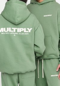 Felpe verde realizzate in morbido tessuto, con grande testo bianco sul retro. Include una tasca a marsupio e dettagli con stringhe sulla cappuccio.