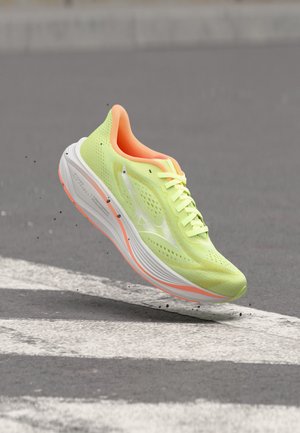 Zapatilla de running de color amarillo brillante con interior naranja y suela blanca mostrada en el aire sobre una carretera pavimentada con marcas viales blancas.