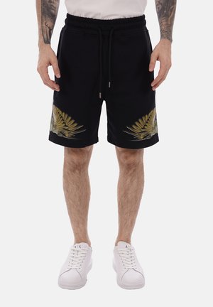 Uomo con pantaloncini neri con design di ali di uccello gialle e verdi vicino all'orlo, scarpe da ginnastica bianche e maglietta bianca, in piedi con le mani rilassate.