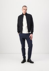 Zwarte gewatteerde bomberjack over een witte gestructureerde sweater, gecombineerd met marineblauwe broeken en zwarte enkellaarsjes. Eenvoudig ontwerp met minimale accenten.