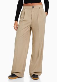 Beżowe spodnie w szerokim kroju z plisami z przodu, z zapięciem na guzik i bocznymi kieszeniami, wykonane z gładkiego materiału, zestawione z czarnymi butami.