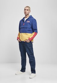 Fargerik lettvektshettejakke med blå, gul og rød seksjoner, med en frontlomme og justerbare mansjetter, kombinert med mørke jeans og joggesko.