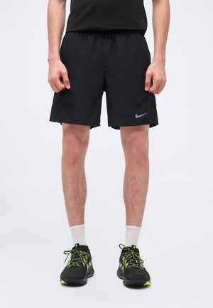 Vyras, dėvintis juodus sportinius šortus su Nike logotipu, baltas kojines ir juodus bėgimo batelius su neoninės žalios spalvos raišteliais ir detalėmis.