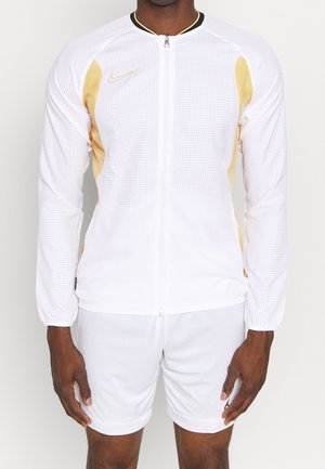 Athlet trägt eine weiße, perforierte Zip-up-Jacke mit goldenen Akzenten und weiße Sportshorts, steht vor einem schlichten Hintergrund.