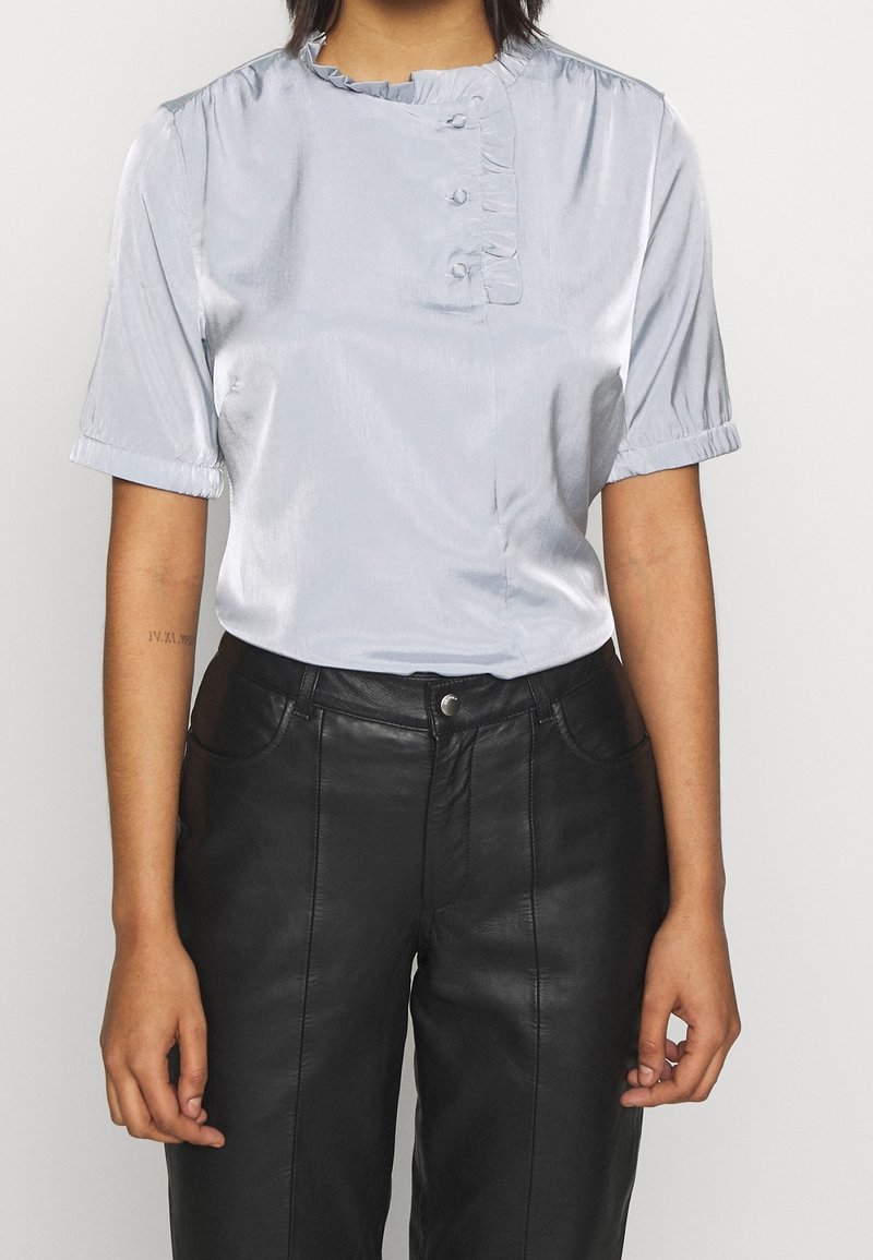 Personne portant un chemisier brillant gris clair à manches courtes avec des volants, associé à un pantalon en cuir noir taille haute.