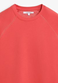 Sudadera color coral con cuello redondo, mangas raglán y puños acanalados. El material parece suave con una textura lisa. Etiqueta de la marca visible.