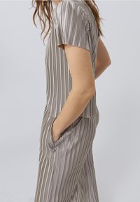 Jumpsuit a rayas gris claro, hecho de un tejido suave y brillante. Cuenta con mangas cortas, bolsillos laterales y un corte holgado. Las líneas verticales crean textura.