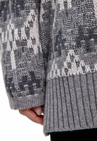 Maglione grigio lavorato a maglia con motivi intricati bianchi e grigio scuro. Orlo a coste, tessuto morbido e maniche lunghe. Mano dettagliata visibile.