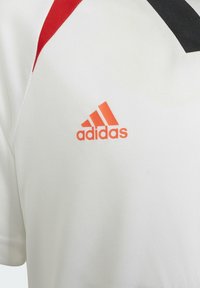 Maillot de sport blanc avec un col V noir, une accent rouge sur l'épaule, et un logo Adidas orange sur la poitrine. Texture du tissu lisse.