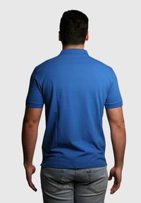 Polo blu a maniche corte, di colore uniforme, tessuto strutturato e colletto classico, abbinato a jeans grigi chiari. Nessun marchio visibile.