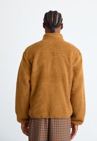 Brown fleece jacket with a high collar, textured surface, and elastic cuffs, worn over checkered pants. Back view showing no visible hardware.