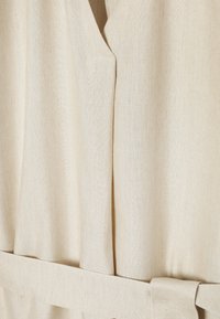 Gros plan d’un vêtement en tissu beige avec une boucle de ceinture et un détail d’encolure en V, montrant un matériau tissé texturé et des coutures.
