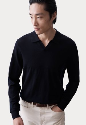 Black long-sleeve polo shirt with a V-neck collar, made of soft fabric, worn with cream-colored pants. The fit appears relaxed.